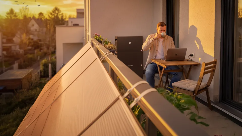 Balkonkraftwerk von Solakon am Geländer eines Stadtbalkons in der Abendsonne, dahinter sitzt ein Mann mit Laptop am Tisch.